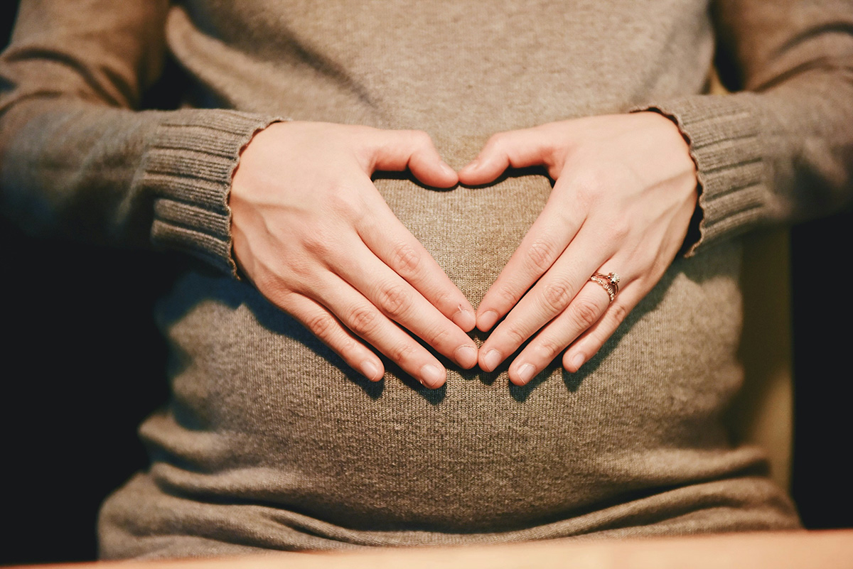 Pregnant woman in tan sweater holding hands over her belly, making a heart shape